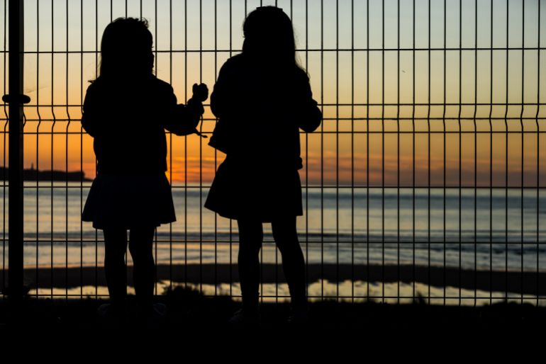 Two backlit girls climbing a metal fence with the orange and yellow fire-colored sky in the background. Concept of relax, sisters, refugee, apocalypse, sadness, poverty, paradise, idyllic, darkness.